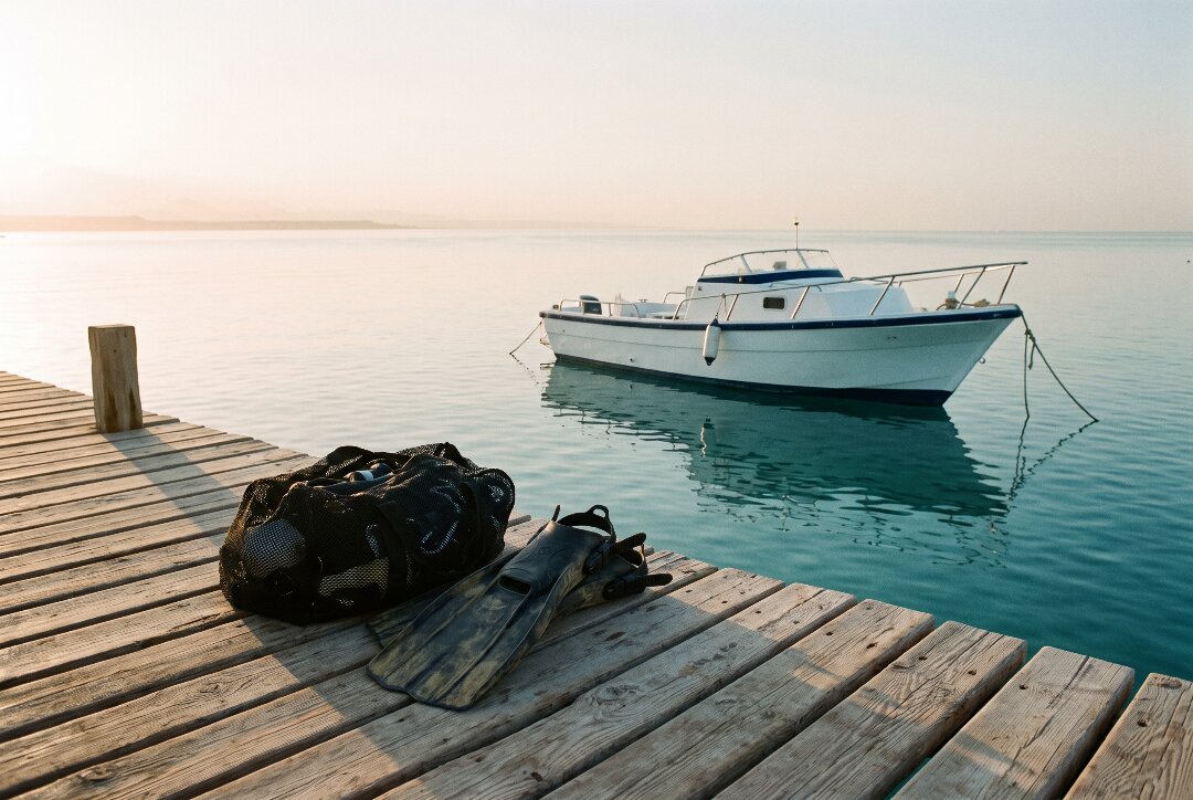 Dive fins and a mesh gear bag on a wooden jetty at dawn, a small boat moored on calm Red Sea water.